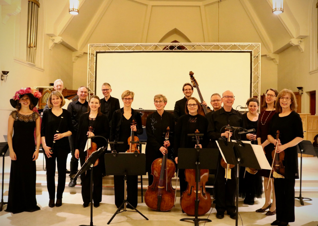 A group photo of the Baroque Odyssey, showing fifteen members smiling holding their instruments on stage.