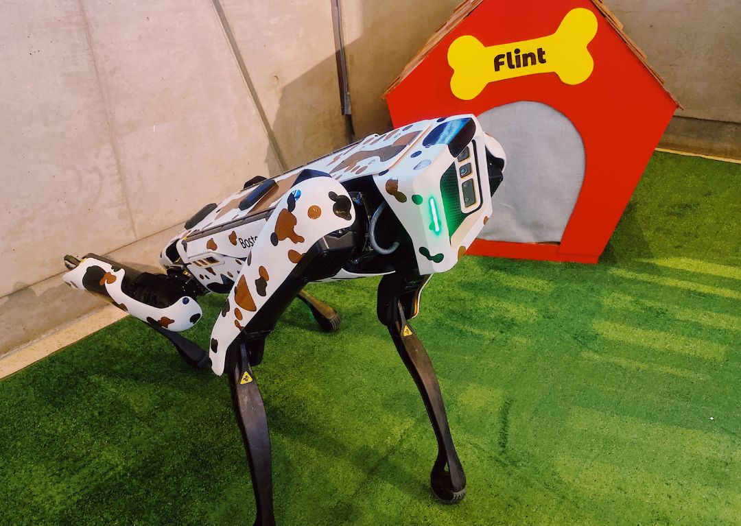 A Boston Dynamics robotic dog sits on astro-turf in front of a dog house.