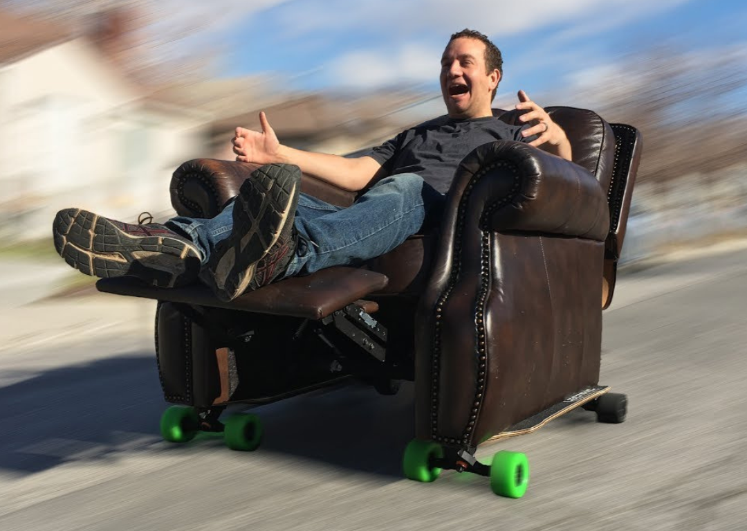 A man smiling as he rides down the street in a sofa chair. The chair is modified with skateboard wheels.