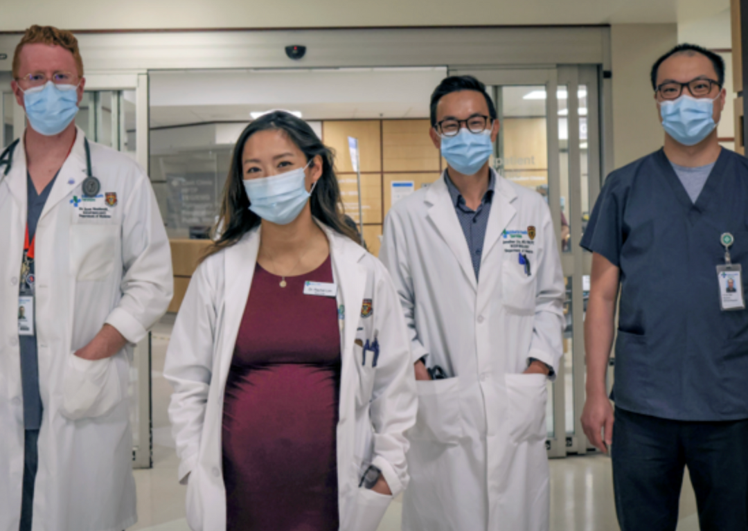 Four doctors walking down a hospital wearing lab coats or scrubs. They are all wearing masks.